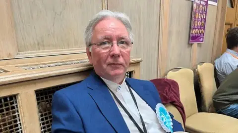 A man with grey hair and glasses in a blue jacket with a white shirt and a light blue rosette sits in front of a wood panelled wall