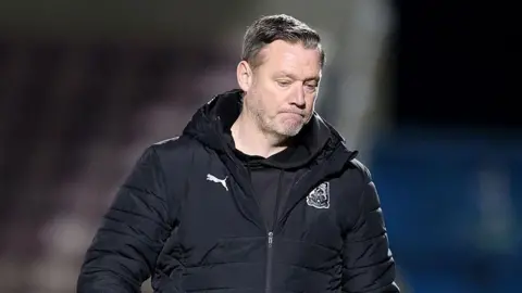 A man in a black sports jacket frowns whilst looking at the floor.