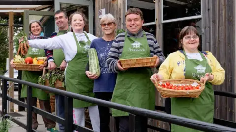 Brunswick Organic Nursery A group of people holding up vegetables, lined up outside a shop and smiling at a camera.