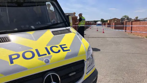 BBC The front of a police van which is parked nearby a police cordon with a police officer. A white forensic tent can be seen in the background.