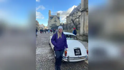 Olesia Sydorenko A woman stands in front of an old-fashioned white car outside a Cambridge University college 