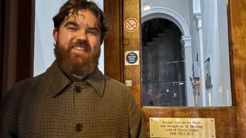 A bearded man, in his 20s, is wearing a smart, brown check jacket. He is is smiling next to a door that displays a golden plaque. It says "Second Star on the Right and Straight on 'til Morning, Land of Green Ginger, Hull, HU1 2EA".