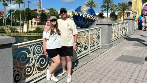 Melissa Rutter A woman with a white t-shirt, black shorts, white trainers, a grey baseball cap and sunglasses standing next to a man wearing a cream t-shirt, black shorts, white trainers, a black baseball cap and sunglasses against white railings, with the Universal Orlando resort in Florida pictured the background.