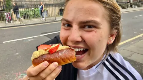 Jasmine is taking a bite out of her cake while looking at the camera. She has short blonde hair and brown eyes. She has a white top on with three black stripes down the shoulders.