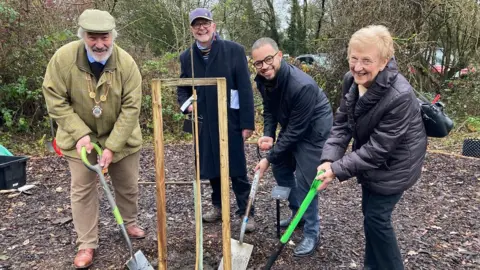 (Left to right) Mayor Robert Bettley-Smith, Colin Bielby from the Minnie Pit commemoration centenary steering group, Adam Jogee Newcastle under Lyme MP and Veronica Billings plant the sapling. They are standing around the tree with spades and smiling at the camera.
