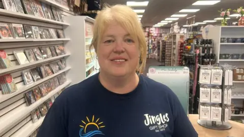Karen McMath has long blonde hair. She is wearing a navy T-shirt with the logo Jingles and a sun logo. A number of pictures and items are visible in the store behind her.
