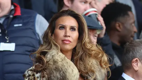 Ampika Pickston has long curled brown hair and is wearing a fur jacket.