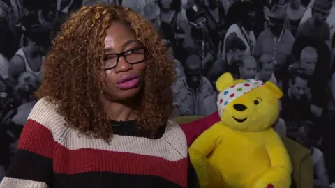 A woman with shoulder-length curly hair wearing glasses and a black, cream and red striped jumper is sitting next to a yellow cuddly bear with a polka dot patch over its eye. It is the BBC Children In Need mascot Pudsey Bear.
