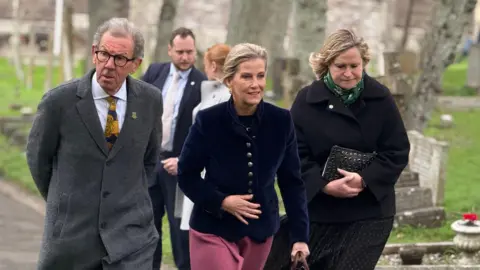 The Duchess of Edinburgh walks through a church yard next to William Tate.