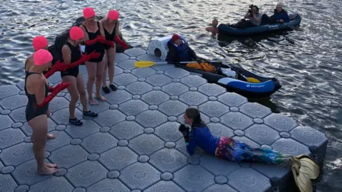 Charlotte Sawyer Woman in a mermaid costume lying on a platform in the water. She is surrounded by five women in black swimsuits, pink swimming caps and red gloves applauding her. In the water there are two paddle boats carrying people, one of which is taking a photo of the scene, and a person is in the water holding on to the paddle boat on the right. 