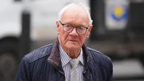 PA Media A man with white hair and black glasses, wearing a navy coat, striped blue and white shirt and striped grey and white tie