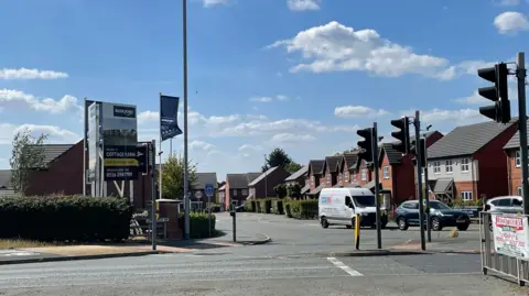 Image shows the entrance to Cottage Farm estate in Oadby. To the left is a sign which says Cottage Farm on it. It is across the road from some traffic lights and there are rows of houses running back on the right. There is a 30mph speed sign in the distance along with a speed bump. 