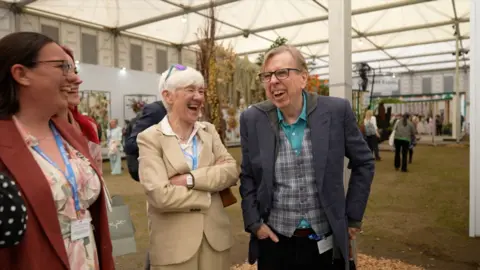 Jonathan Swingler/BBC Timothy Spall laughing. He has light, short hair and is wearing dark glasses, a blue blazer, tartan waistcoat and lighter blue shirt underneath. One of his hands is in his trouser pockets while the other rests by his side. Three women are standing to the left also laughing. The one closest to him has white, short hair and is wearing a beige suit. There is a woman with red hair next to her, but she is mostly blocked by a woman with dark hair and glasses. They are standing where the exhibitions are which is a large marquee with grass on the floor.