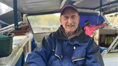 A man wearing a blue baseball cap, a tartan scarf and a blue waterproof jacket sitting on a boat. The interior of the boat and a window with a view over the river can be seen behind him.