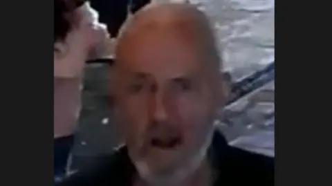 A grainy CCTV image of a bald man with a white short beard in a dark polo shirt. 