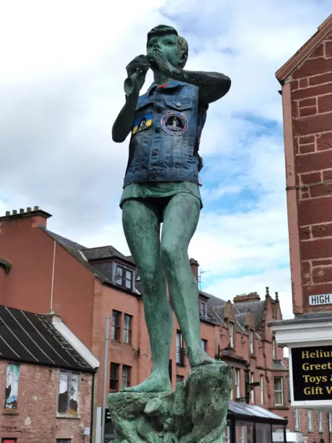 Alex Macleod A Peter Pan statue wearing a cut-off denim jacket in a town square