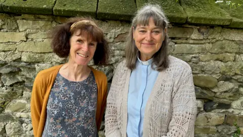 Sarah Lister Two older women stand side by side, smiling. The woman on the left has short brown hair held back with a headband, and wears a blue floral patterned top and a mustard coloured cardigan. The woman on the left wears a vicar's collar, a blue shirt and a beige cardigan.