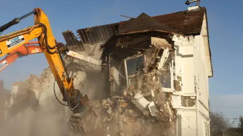 PA Media A yellow and orange digger knocking down a white house with a brown roof.