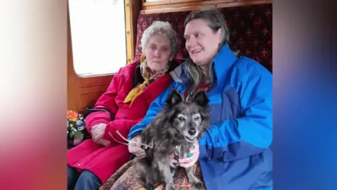 Phazar Souter Miriam Souter has short grey hair and is wearing a pink coat and yellow scarf. Sat next to her is her daughter Phazar, who has platted brown hair and is wearing a blue coat. On Phazar's lap is a medium sized black dog.