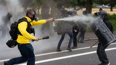 Getty Images Członkowie dumnych chłopców i Antifa Clash w Portland w stanie Oregon. Mężczyzna ubrany w maskę gazową i żółtą kurtkę rozpyla kanister w kierunku czarnego odzianego mężczyzny trzymającego tarczę. 