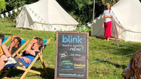 Two bell tents and people laying on deck chairs next to a sign which asks, 'need a space to relax?'