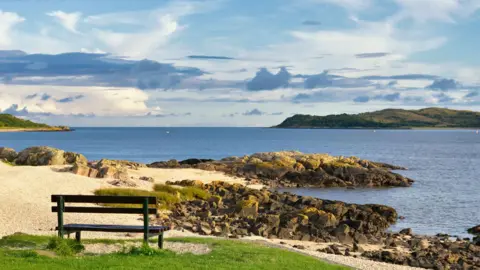 Getty Images A bench looking out to sea across the Solway Firth with rocks jutting out into the water and green hills on the horizon