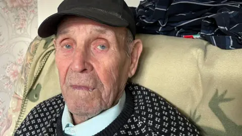 George Brown, 92, has a black peaked cap on and small tufts of grey hair are visible on the sides he is wearing a brown and white patterned jumper with a light blue collared shirt underneath