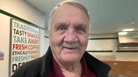 BBC/Louise Hobson Mick Male is a man with short grey hair, a red top and a black fleece. He is stood in a large room.