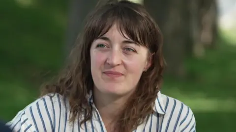 Clinical psychologist Dr Ruth Denton, a woman with brown shoulder-length wavy hair and wearing a blue and white striped shirt, sits on a park bench. The background is out of focus but green shrubbery and trees can be made out.