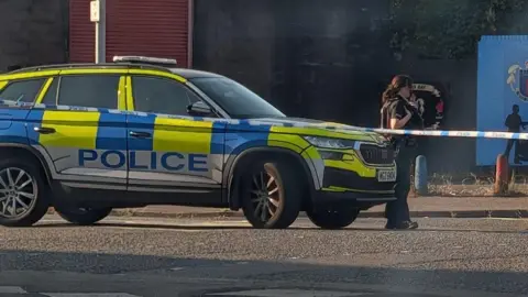 A police officer is behind tape that's cordoning off a road. A police car is parked. A mural saying loyalist Tigersbay is on a wall that's been painted blue.

