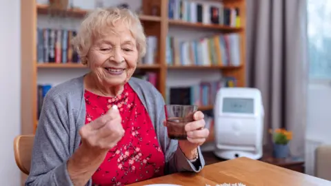 An elderly woman wearing a red floral dress and grey cardigan. She is holding a glass of water and a pill. She is sat at a wooden table. Behind her is a white wall and bookshelf. 