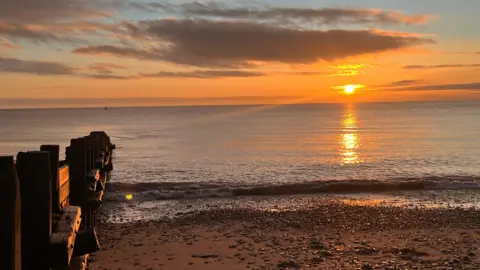 A sunset over a calm sea with orange and gold reflections on the water. A pebble beach is visible in the foreground, and wooden groynes extend into the shoreline.