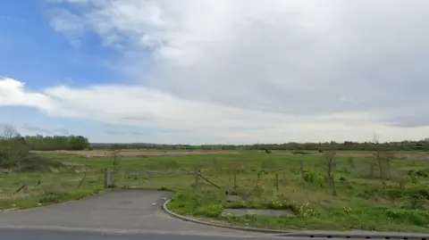 Google An empty grass plain with a gate and path in the foreground