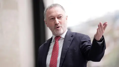 Steve Reed, a man in his late fifties with short grey hair and a white beard, speaking and looking to the right wearing a suit and red tie pointing his left arm towards the camera.