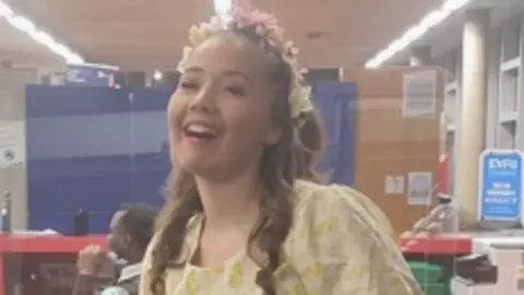 Northamptonshire Police A blurry close up photo of Ella Walden, wearing a yellow and pink flowers in her long curling mid brown hair. She is wearing a yellow and white dress and is in a large room. A man can be seen sitting behind her, turned away. 