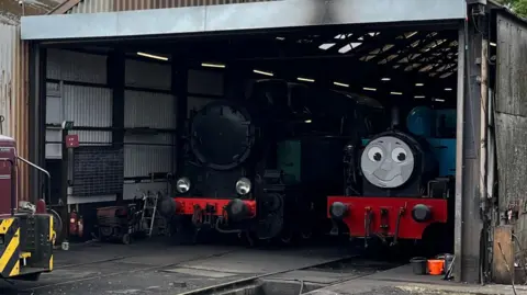 Submitted A close-up of Thomas a locomotive at Nene Valley Railway. The blue locomotive has a smiling face painted on the front of its engine. It is sitting in a shed in sidings. 