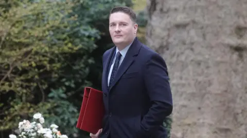 Streeting walking outdoors in what appears to be a landscaped garden or park area. He is wearing a long, dark overcoat and holding a bright red folder under one arm. The background features dense greenery and flowers.
