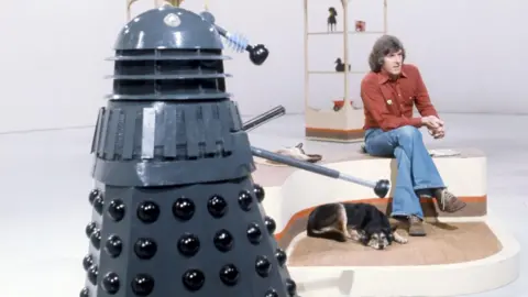 A colour photo of a 1970s television studio, with a primarily white background and floor, with some free-standing shelving in the background. Dominating the photo close up at the left of frame is a Dalek, a conical machine creature, dark grey in colour with black trim. On its right, looking away to a camera out-of-frame, is a man in his late 30s with long, greying dark hair, wearing a red shirt and flared blue jeans. At his feet is a slumbering black-and-brown dog.