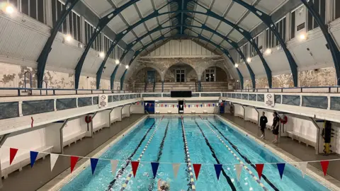 A view from a balcony of the pool, which stretches out into the distance. It has a large vaulted roof above it with arched metal beams, There is traditional brickwork and tilting as well as bunting hung above the pool