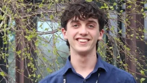 Lucas smiling at the camera with a tree behind him. He's wearing a blue shirt. 