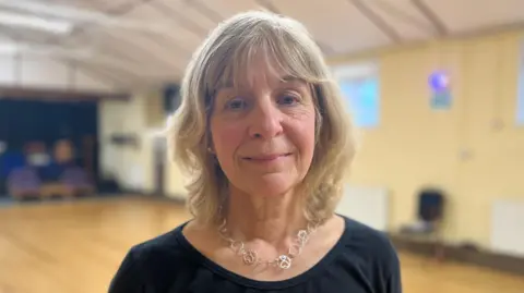 Aimee Dexter/BBC Audrey Cater is standing in a hall. She has shoulder length blonde hair and is wearing a black T-shirt and a silver necklace.