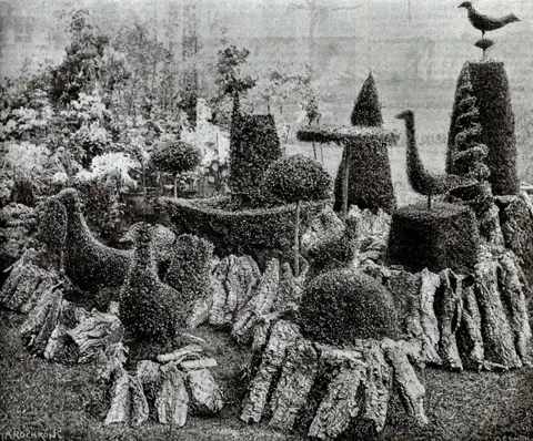 RHS Lindley Collections a photo of some topiary in different shapes set out in a circle. The topiary bushes are shaped into birds, circular mounds, vertical spirals and there is a wheelbarrow one in the middle. They are positioned on top of what looks like sawn off tree trunks.