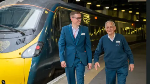 PA Two railway staff in uniform walk along a station platform next to a high‑speed train under overhead lights.