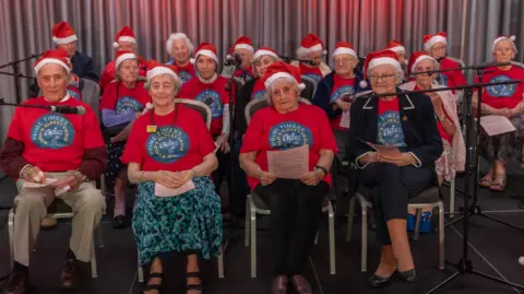 A number of elderly people wearing red shirts and Santa hats sitting on chairs upon a stage. Microphones have been placed on either side of them. A silver curtain is visible in the background.