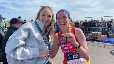 Apryl Hammett Apryl and Paula Radcliffe smile for the camera at the Brighton Marathon. Apryl in a red vest, which says Breast Cancer Now on the front and Paula in a grey tracksuit top.