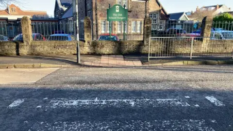 A faded zebra crossing stretches from the centre of the foreground to the centre of the shot. A green sign reading "The Old School Surgery" is in the centre background, with railing either side of it.