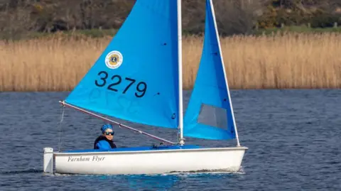 Kim Davies A woman in a white boat with a blue sail on the water.