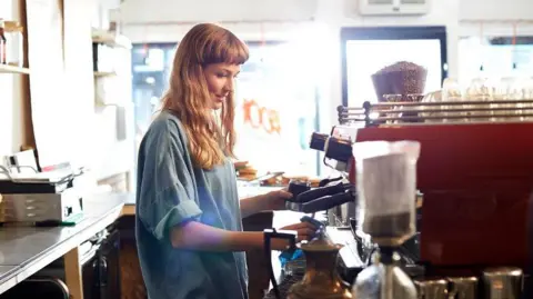 A barista in a coffee shop, she is wearing a blue denim shirt and making a coffee at a red coffee machine. She has long red hair with a blunt fringe. 