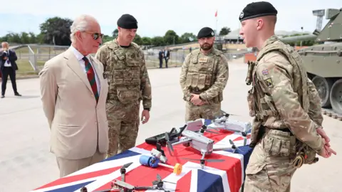PA Media King Charles in a cream coloured suit with a red and green tie and sunglasses. He is talking to three men dressed in camouflaged military uniform, with a table full of drones, covered in a British flag, in between them. There is a tank in the background.