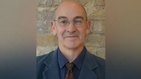 Amy Holmes/BBC Head and shoulders image of Ben Crystall in front of a brick wall. He is wearing glasses, a dark blue jacket and shirt and a brown tie.